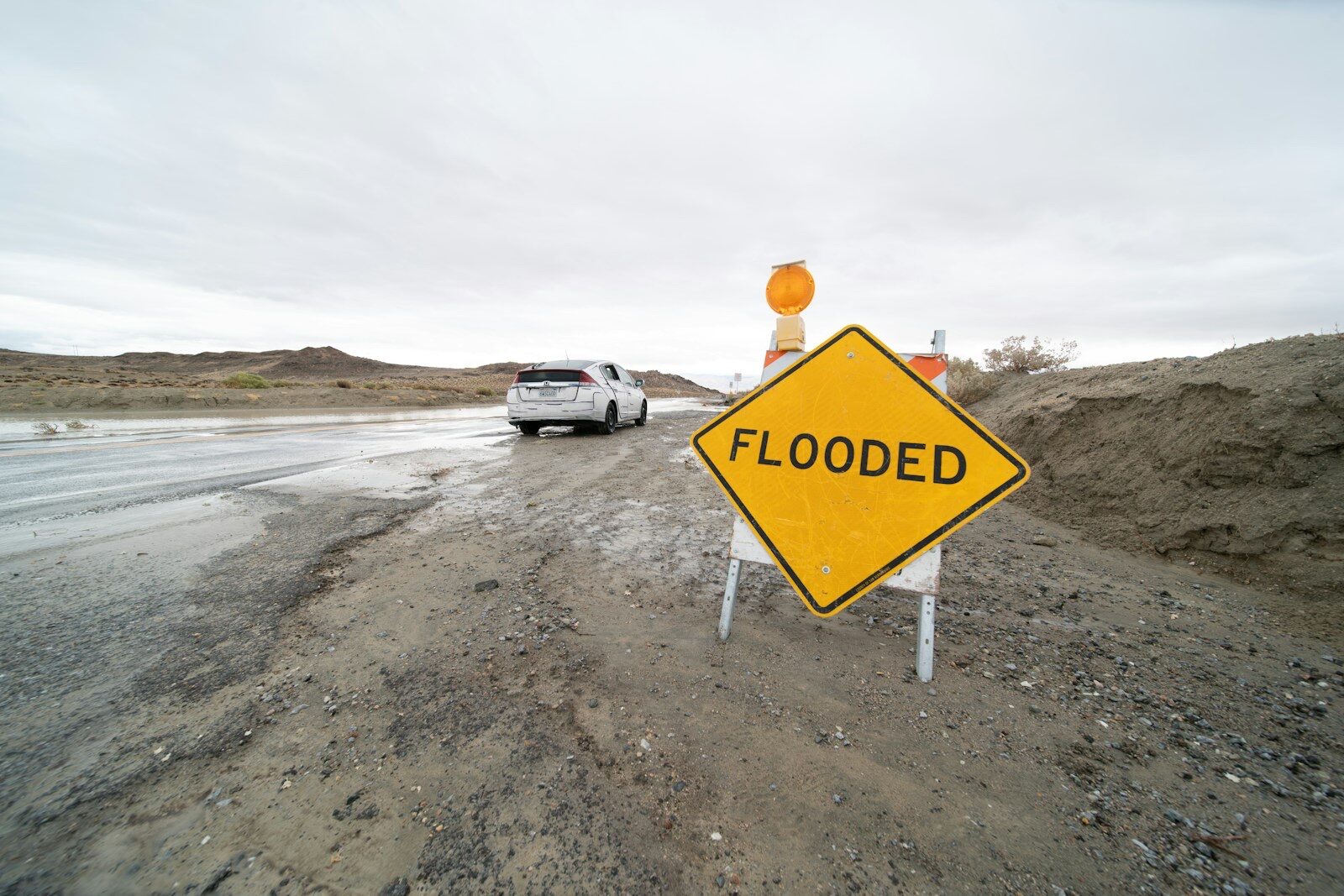 A yellow diamond road sign reading FLOODED stands on muddy gravel beside a desert road running with water, as a white car drives through under a grey sky.