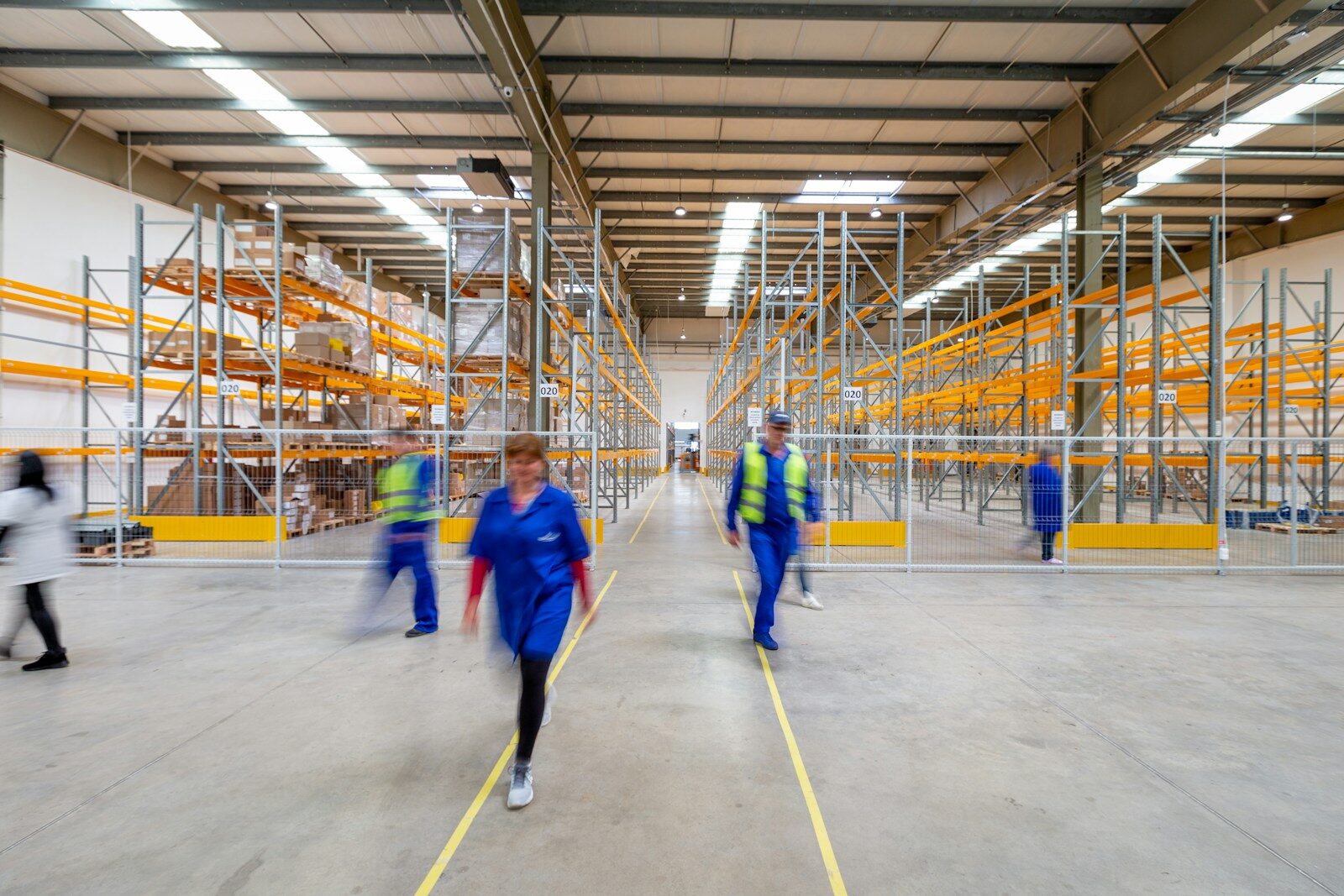 Workers walking through a large distribution warehouse lined with pallet racking, captured with motion blur.