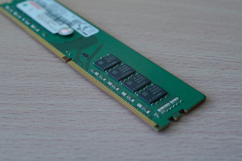 A green computer memory module with a row of black memory chips lies on a light wooden surface, photographed close up with a shallow depth of field.
