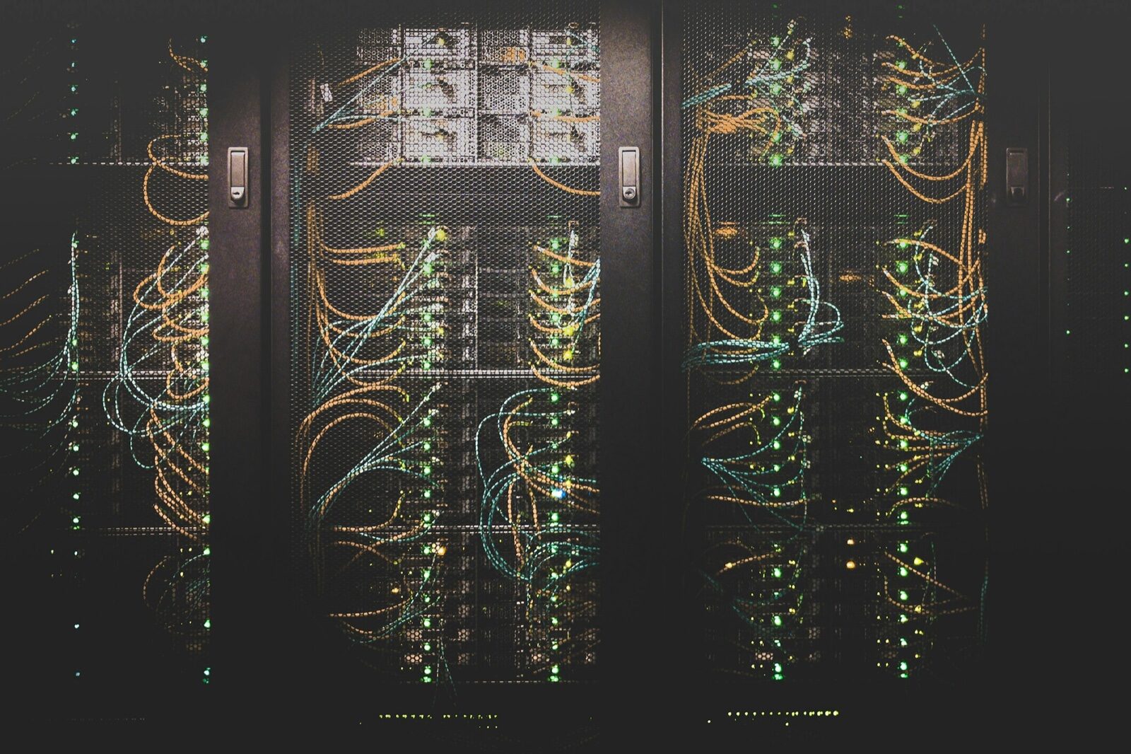 Three server racks in a dark room behind mesh doors, threaded with orange and blue network cables and dotted with green status lights.
