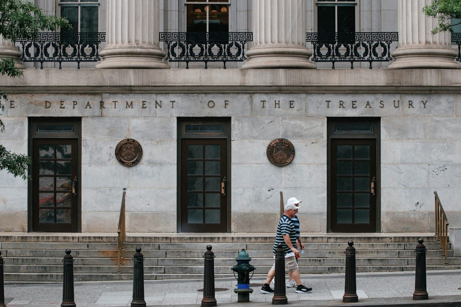 Two men walk past the stone facade of the Treasury Department building in Washington, with 'The Department of the Treasury' carved above three doors and seals mounted between them.