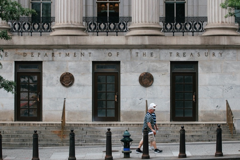 Two men walk past the stone facade of the Treasury Department building in Washington, with 'The Department of the Treasury' carved above three doors and seals mounted between them.