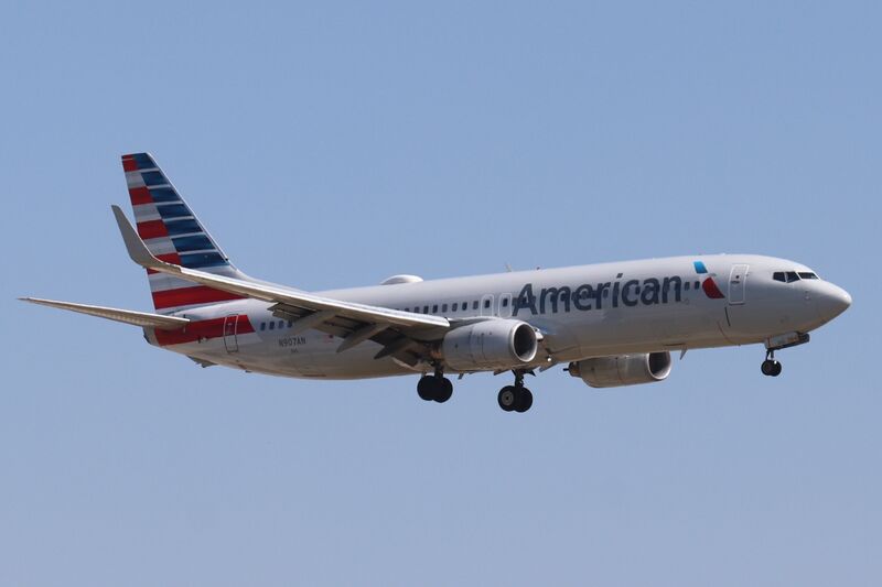 An American Airlines Boeing 737-800 in silver livery with a striped red, white and blue tail, landing gear down, on approach against a clear blue sky.