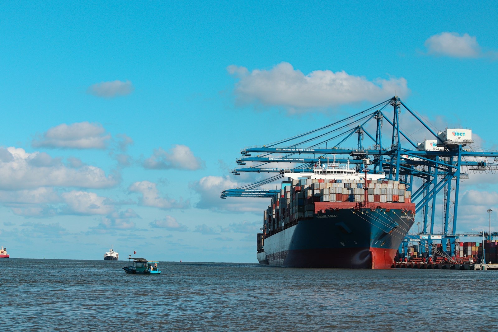 A cargo ship loaded with stacked shipping containers sits beside blue port cranes under a blue sky with scattered clouds, with a small boat on the water in front.