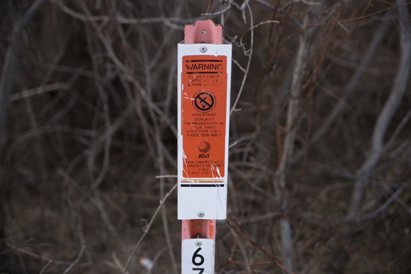 An orange AT&T warning sign on a post among bare branches, reading Warning, buried fiber optic cable in this vicinity.