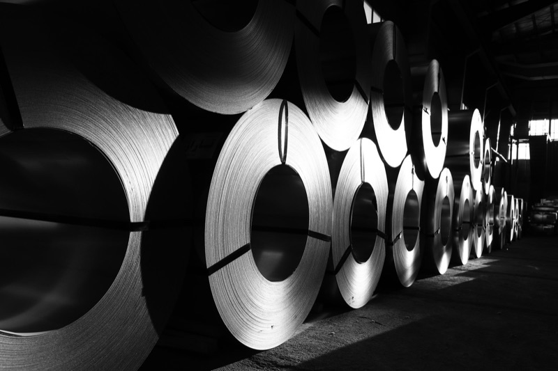 Black-and-white photograph of large rolls of sheet steel standing on their edges in a long row inside a dim warehouse, lit by a shaft of light from the right.