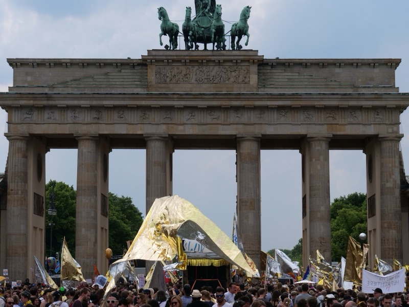 A large crowd with flags and a gold-foil float gathered in front of the Brandenburg Gate.