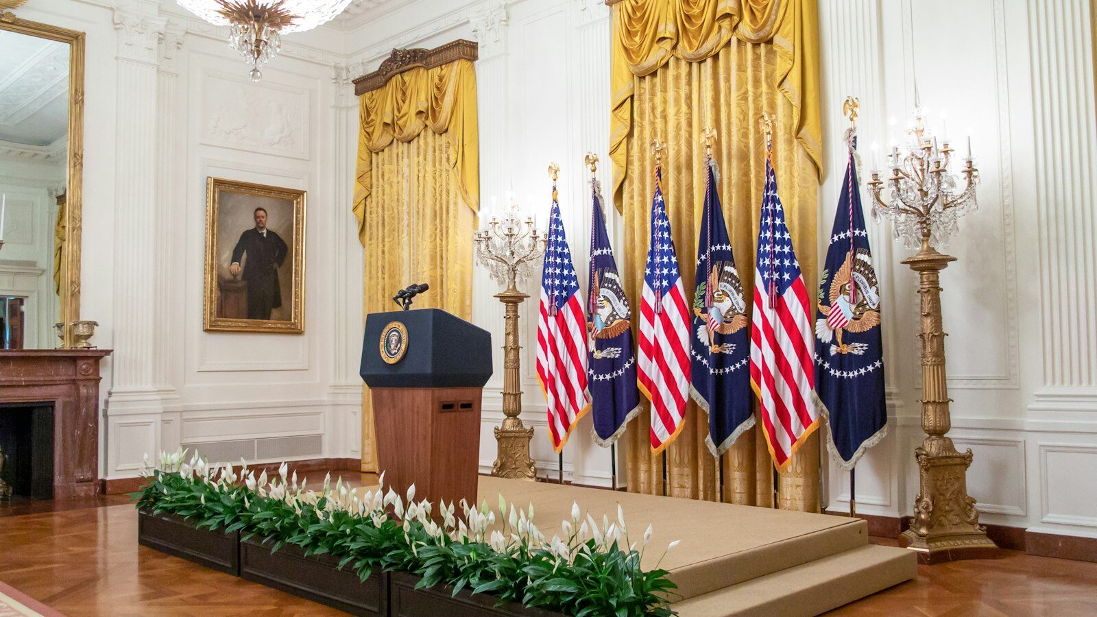The empty presidential podium in the East Room of the White House, flanked by flags and a portrait of Theodore Roosevelt.