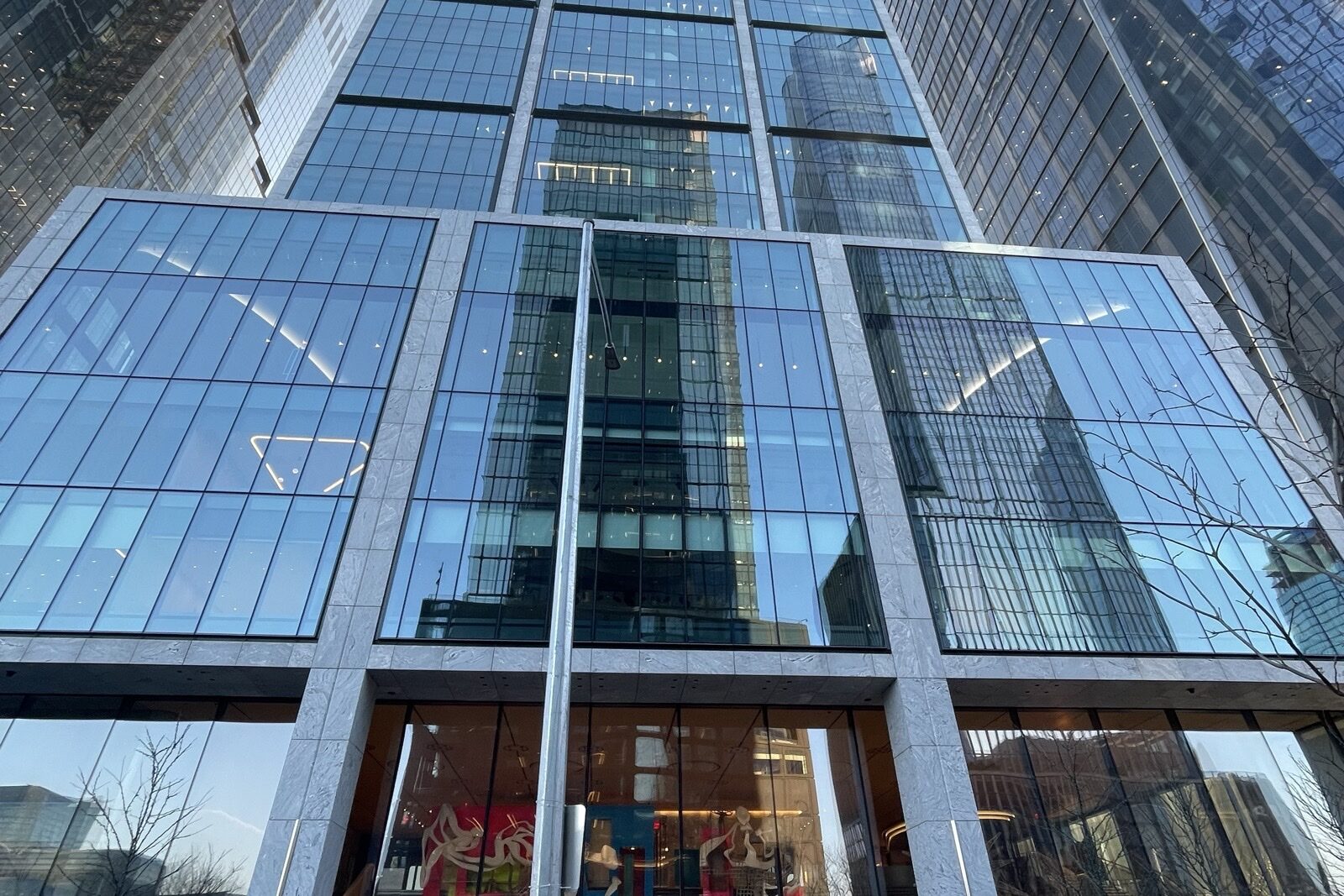 The glass facade and stone-framed lower floors of 50 Hudson Yards in Manhattan, seen from the street, with neighbouring towers reflected in the windows.