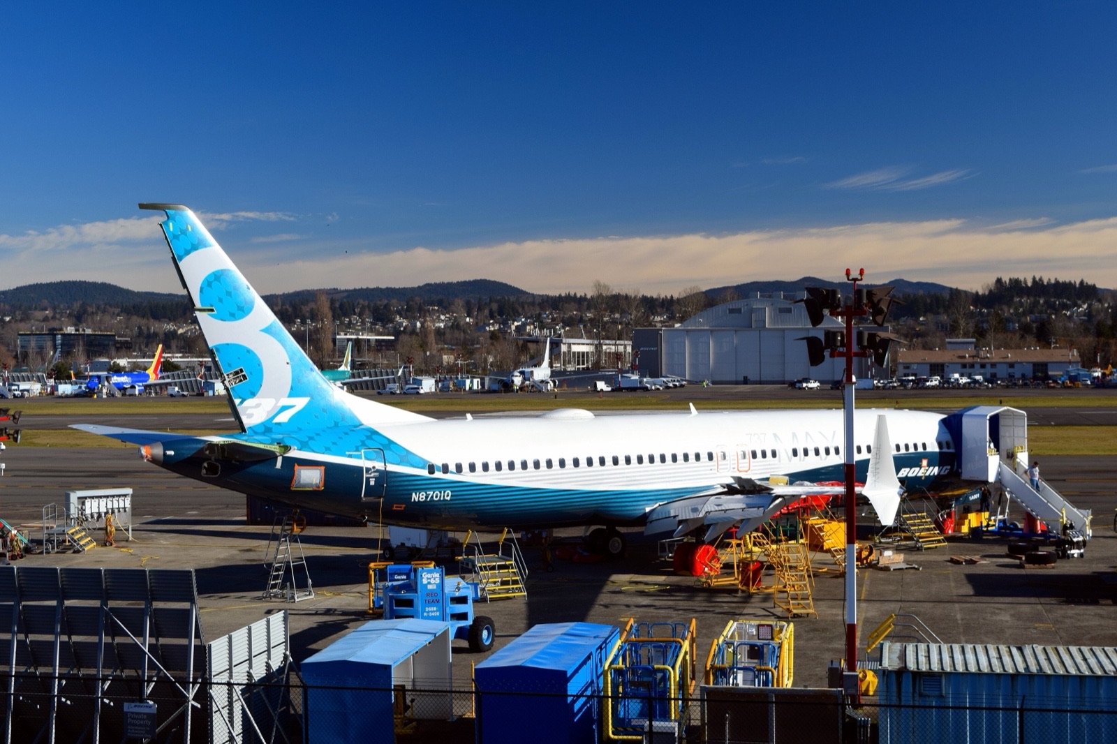 A Boeing 737 MAX test aircraft in Boeing's blue and white livery, registration N8701Q, parked on an apron with work stands and stairs beside it, with hangars, hills and a blue sky behind.