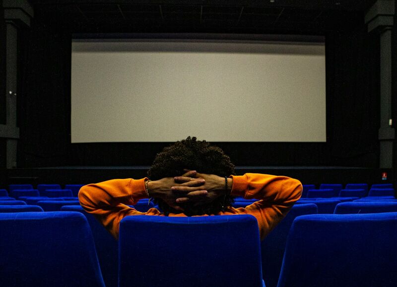 A man in an orange sweatshirt sits alone in a cinema's blue seats, hands clasped behind his head, facing a blank screen.