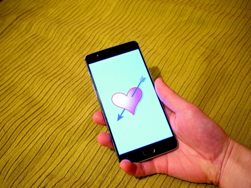 A hand holds a smartphone whose screen shows a pink heart pierced by an arrow, against a green textured fabric.