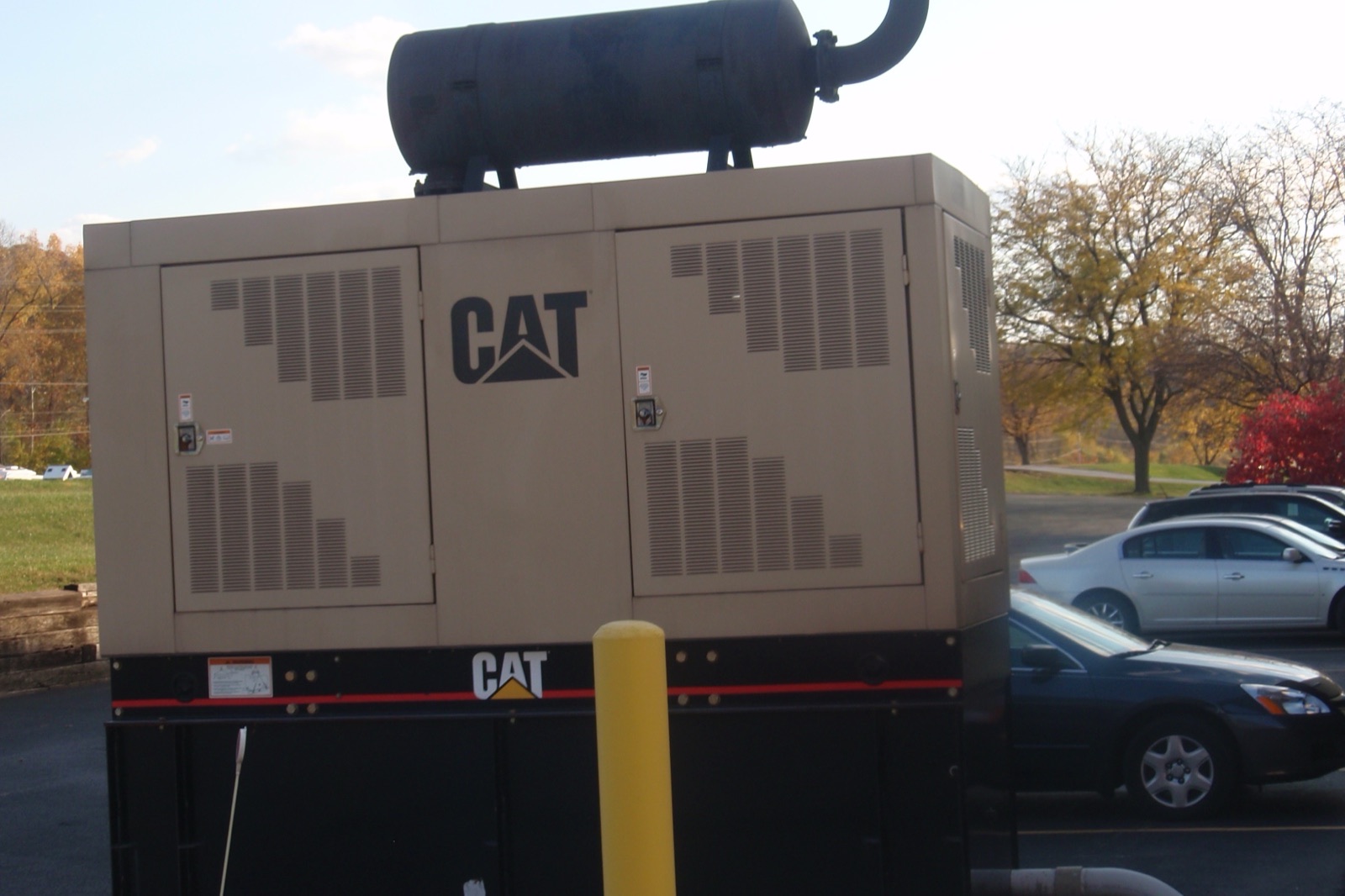 A beige diesel generator set with CAT lettering on its enclosure and an exhaust silencer on top stands in a parking lot beside parked cars, with autumn trees behind.