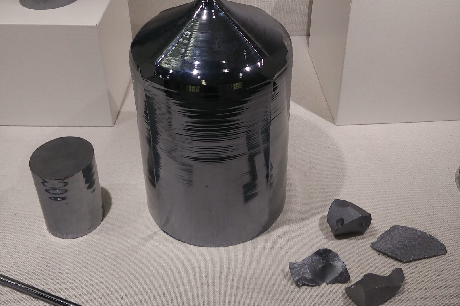 A polished grey monocrystalline silicon ingot on a museum display plinth, with a smaller cut silicon cylinder to its left and rough chunks of silicon in front of it.
