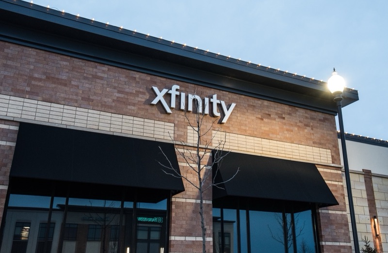 The front of an Xfinity store in a brick retail building at dusk, with the white Xfinity sign above two black awnings, a bare young tree and a lit street lamp.