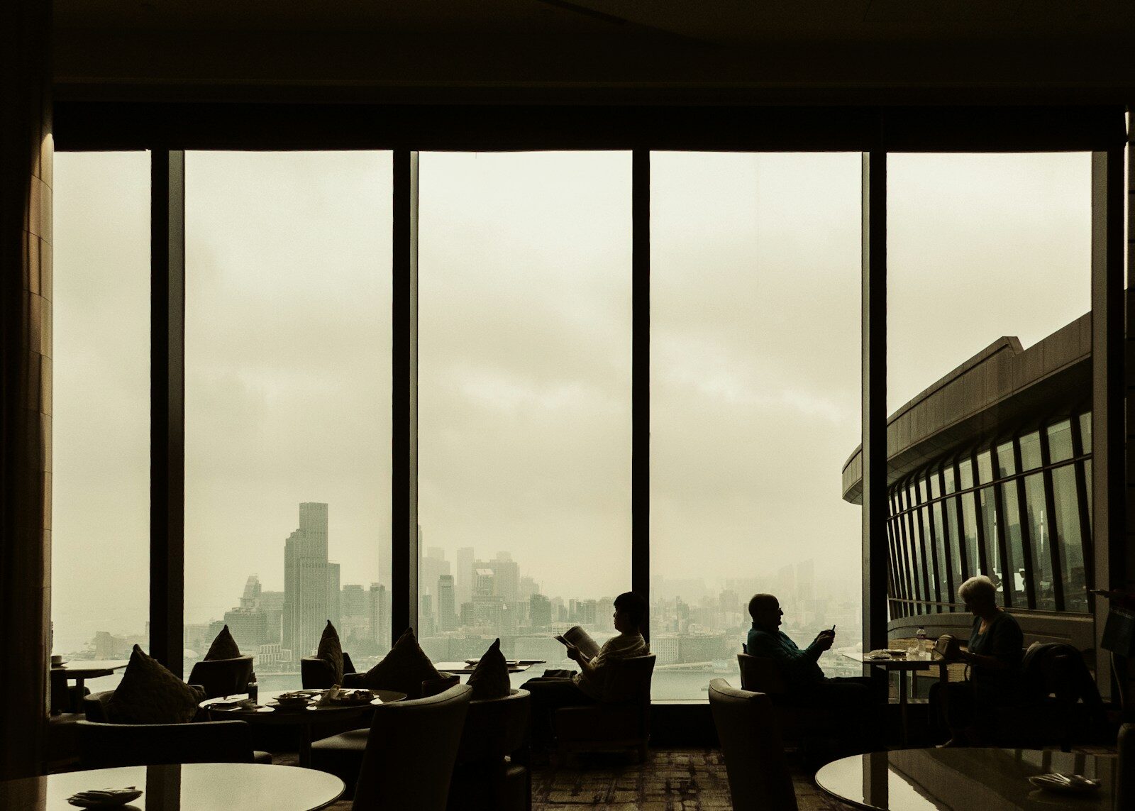 Silhouetted figures relax in the serene, dimly lit lounge of a high-floor Hong Kong Island hotel. Through expansive floor-to-ceiling windows, the iconic Kowloon skyline emerges from a thick, atmospheric mist, creating a soft, monochromatic panorama. People enjoy quiet, personal moments against the vast cityscape, a tranquil sanctuary high above the metropolis.