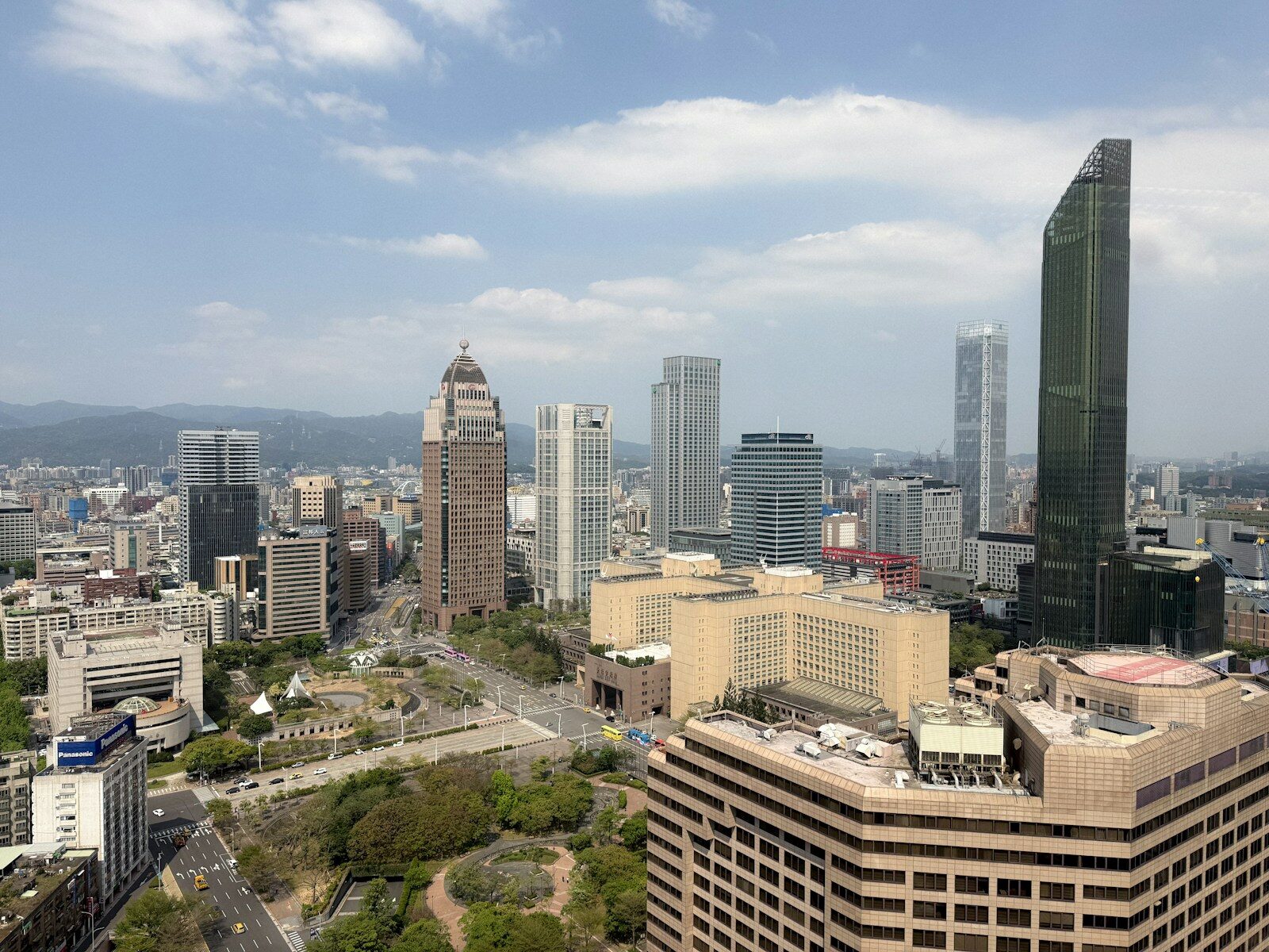 Aerial view of Taipei's Xinyi district skyline from a high-rise, showing office towers, a broad boulevard, and green park space beneath a partly cloudy sky.