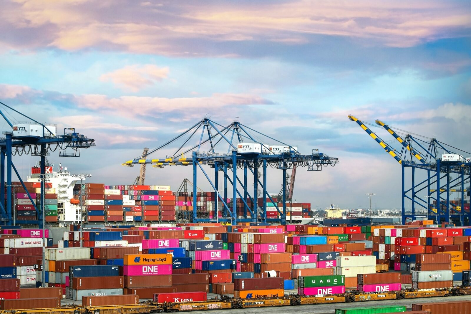 Stacks of shipping containers from several lines at a container terminal in the Port of Los Angeles, with blue ship-to-shore cranes against a pink and blue evening sky.