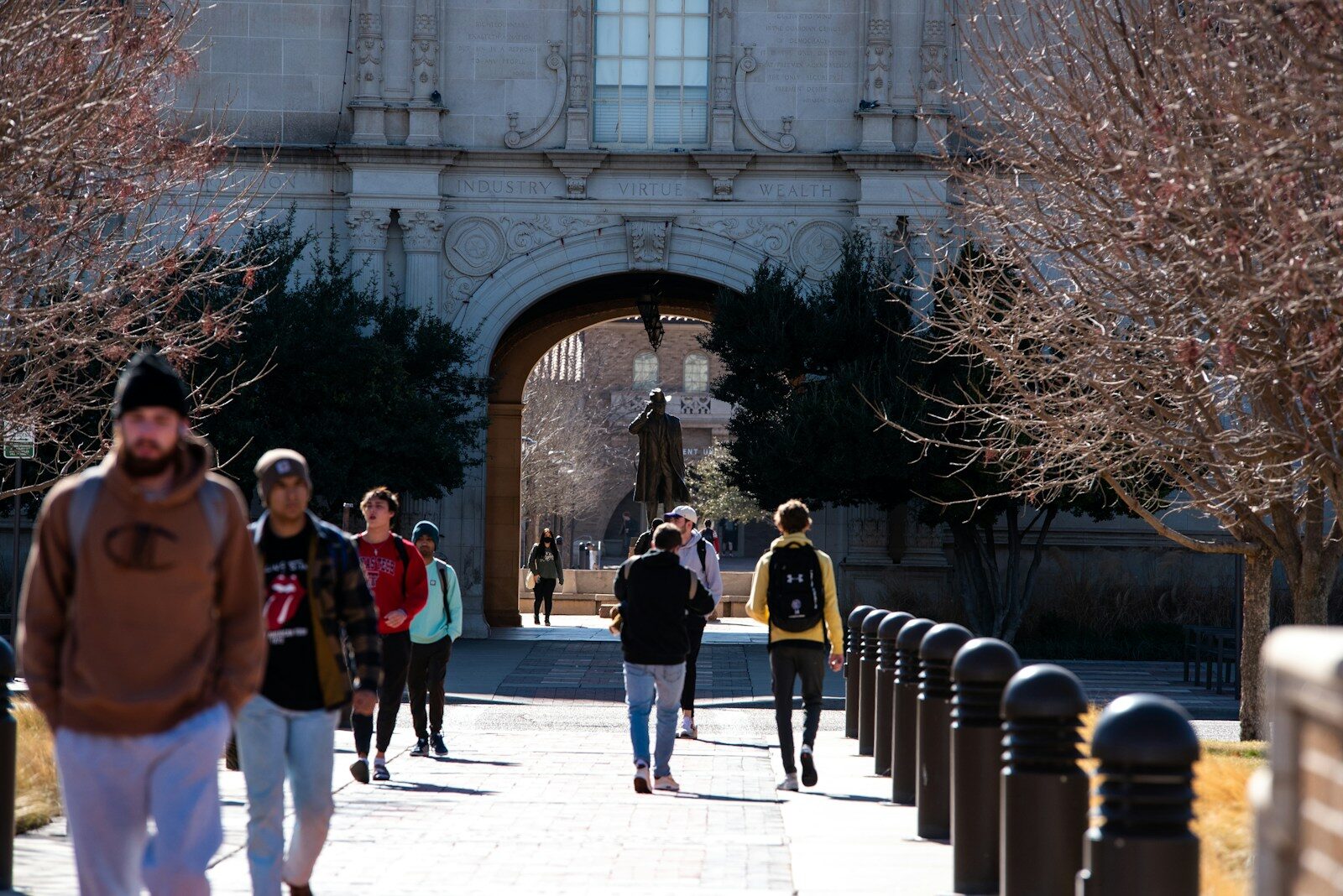 Students walking on a campus path towards a stone archway inscribed INDUSTRY VIRTUE WEALTH.