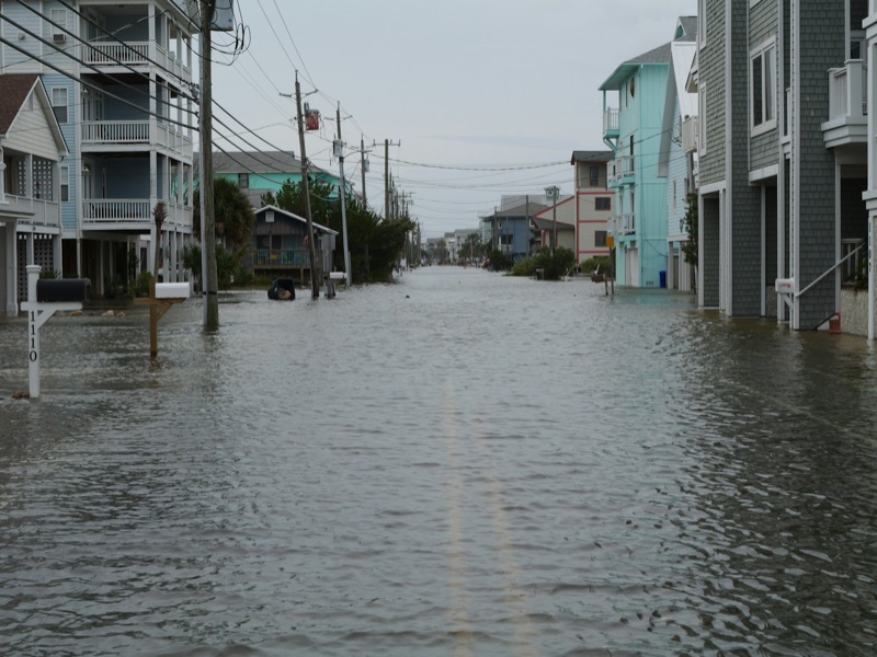A coastal residential street under a foot or more of standing water, the centre line just visible beneath the surface, with stilted beach houses, mailboxes and utility poles on both sides under a flat grey sky.