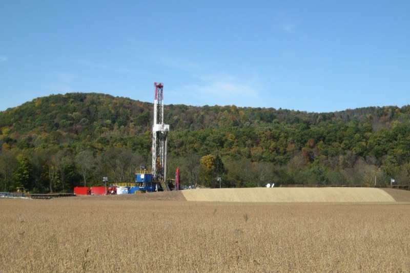 A tall drilling rig with a red-and-white mast stands on a levelled gravel pad at the foot of a wooded hill in autumn colours, seen across a field of ripe brown soybeans under a clear blue sky.