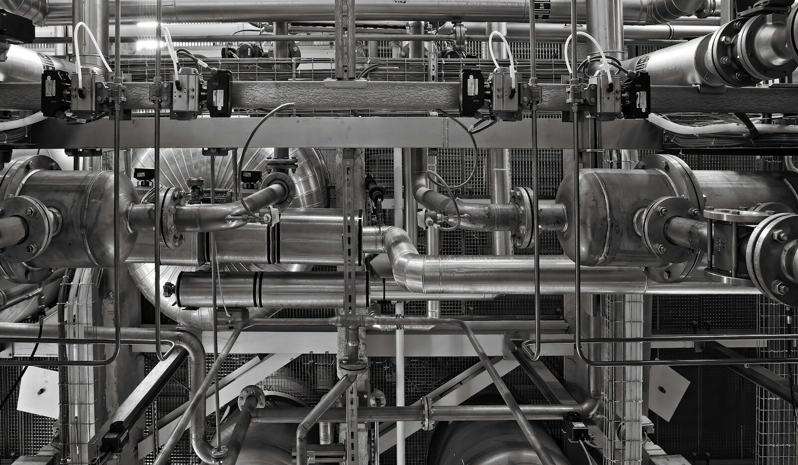 Stainless-steel pipes, valves and steel framing inside an industrial plant, in black and white.