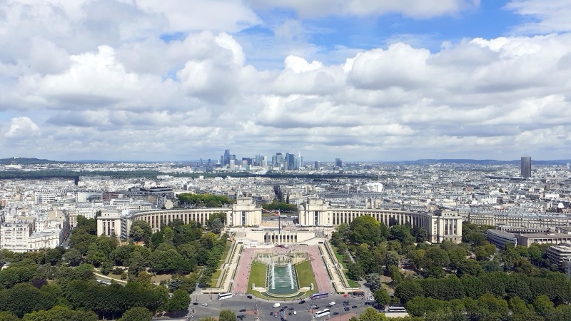 A view over Paris from the Eiffel Tower, looking across the Trocadéro gardens and the curved Palais de Chaillot towards the towers of La Défense on the horizon under heavy white clouds.