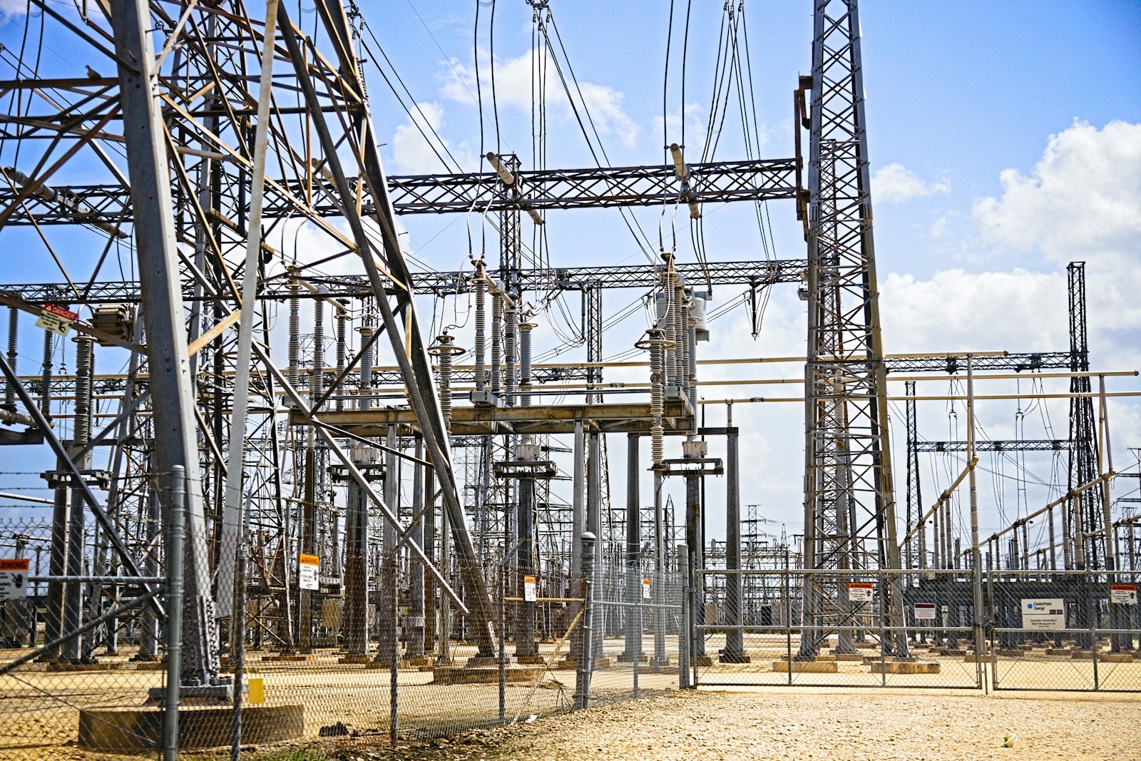 A dense high-voltage electricity substation, steel gantries and insulators crowded behind a perimeter fence.