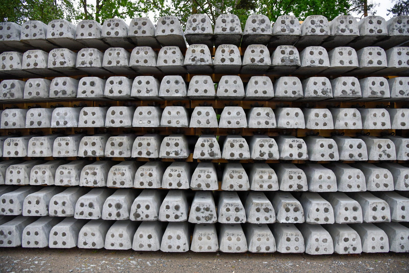 Rows of concrete railway sleepers stacked several pallets high, awaiting installation beside a treeline.