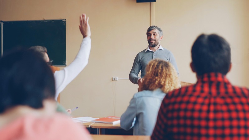 A teacher addressing a class, with a student raising a hand.