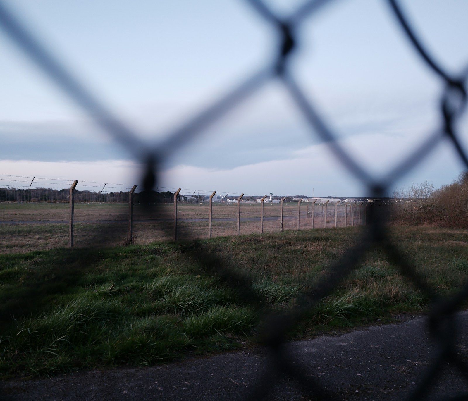 A chain-link fence in the foreground, out of focus, with a second post-and-wire fence running across flat grassland beyond it towards a line of low buildings and trees on the horizon under an overcast sky. A strip of tarmac runs along the bottom edge. No people, vehicles or aircraft are visible.