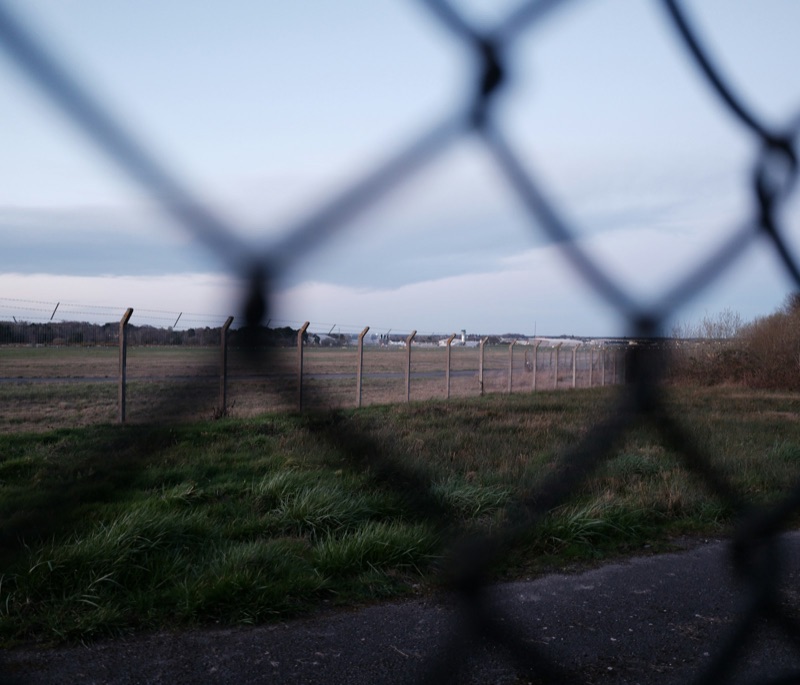 A chain-link fence in the foreground, out of focus, with a second post-and-wire fence running across flat grassland beyond it towards a line of low buildings and trees on the horizon under an overcast sky. A strip of tarmac runs along the bottom edge. No people, vehicles or aircraft are visible.