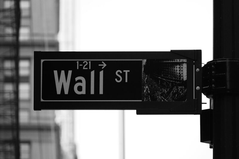 A Wall St street sign on a lamp post in black and white, with office buildings blurred behind it.