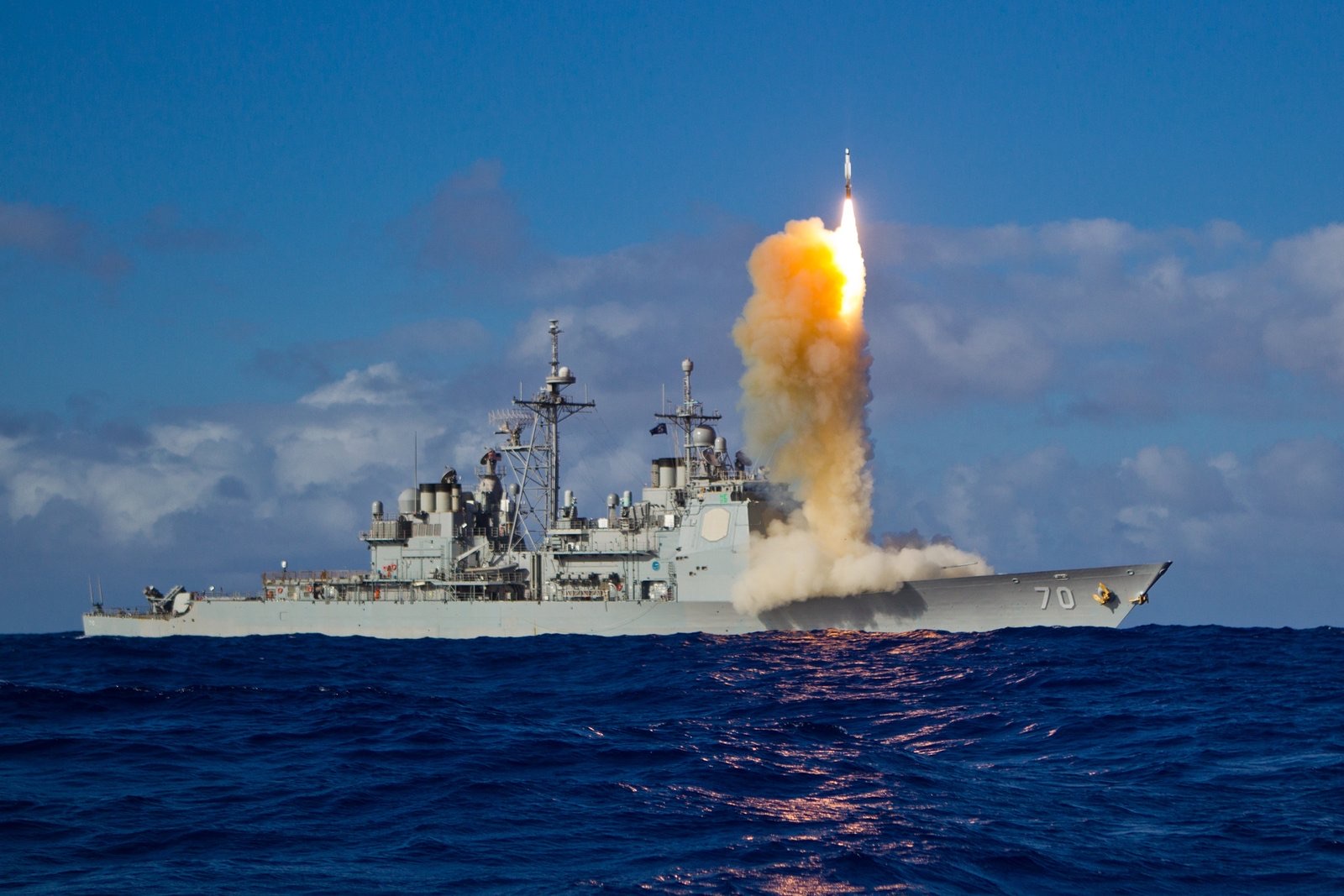 A grey Navy cruiser with hull number 70 at sea under a blue sky, a missile rising from its forward deck on a column of orange flame and billowing smoke.