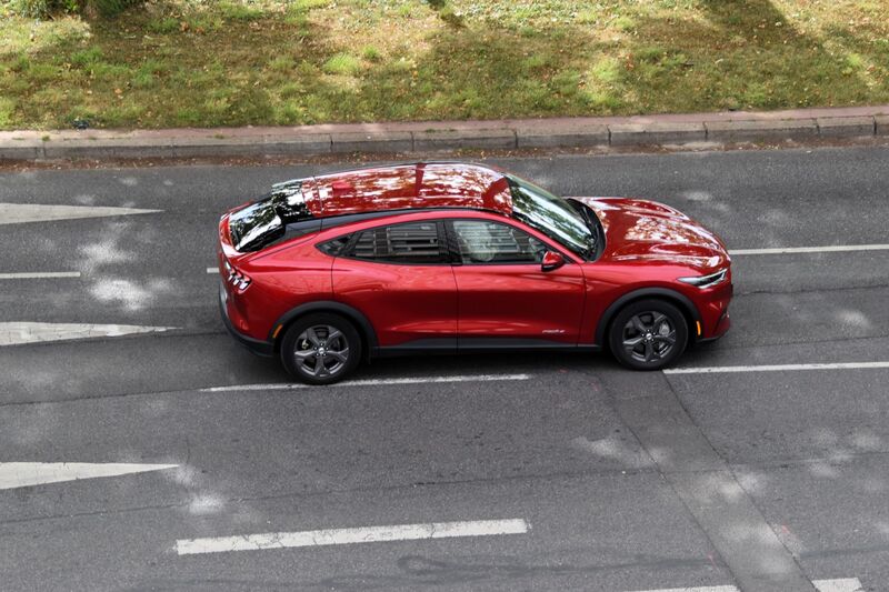 A red Ford Mustang Mach-E electric crossover seen from above and the side, driving along a grey road with white lane markings beside a grass verge.