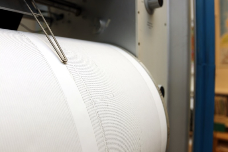 A close-up of a white seismograph drum wrapped in paper ruled with fine recorded lines, with a thin metal stylus resting on it.