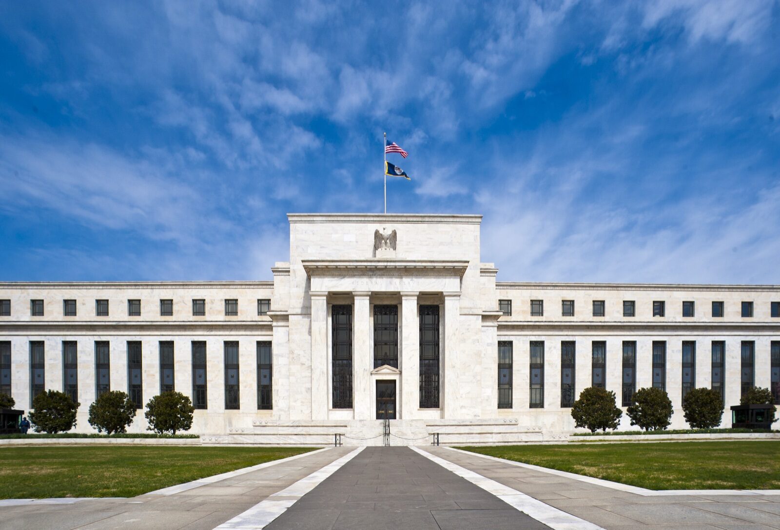 The white marble facade of the Federal Reserve's Eccles Building in Washington under a blue sky, with an American flag above the entrance.