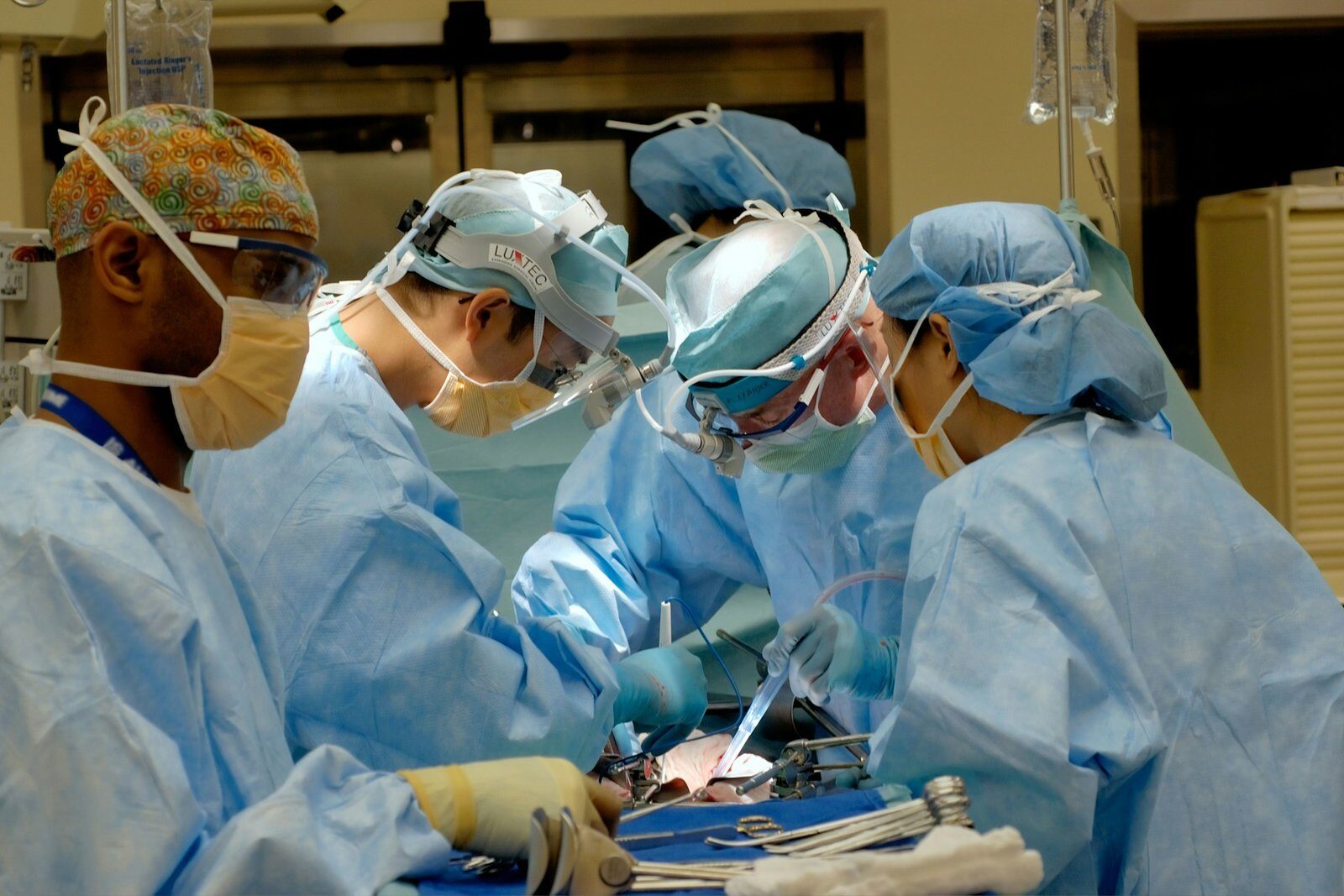 Four surgeons in blue gowns, caps and masks, two wearing magnifying headlamps, lean over a patient during an operation in a brightly lit operating room.