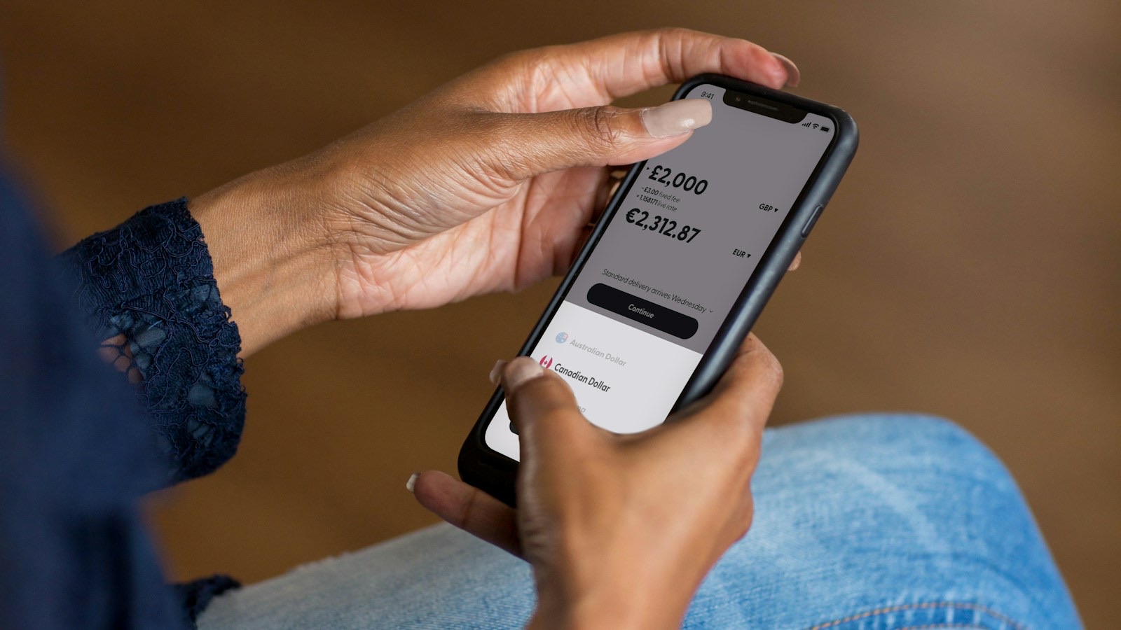 A pair of hands holding a smartphone, the screen showing a banking app with a currency amount and a transfer confirmation button.