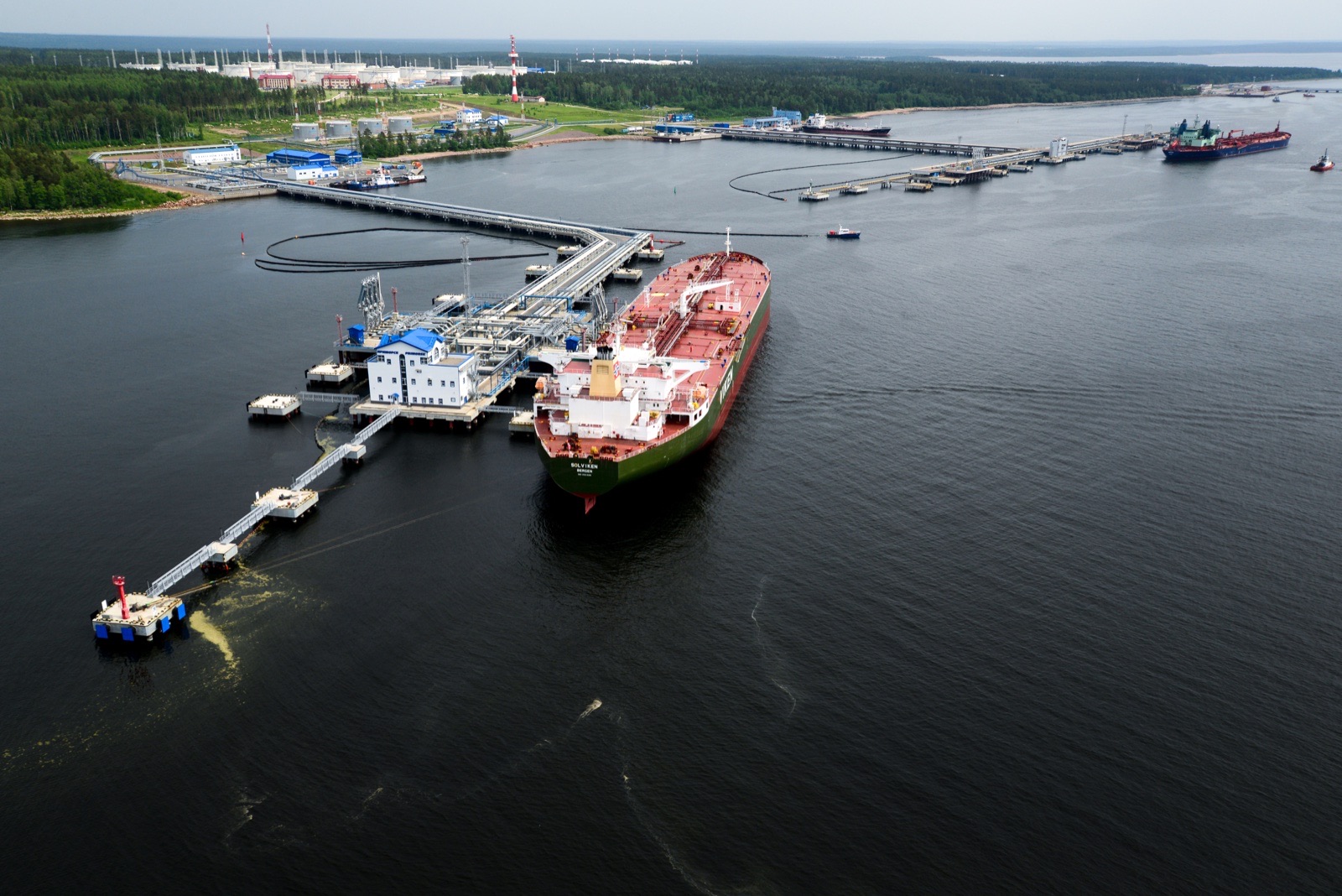 An aerial view of a large tanker with a green hull and red deck moored at a jetty of the Primorsk oil terminal, with a second tanker at a berth in the distance and forest along the shore.