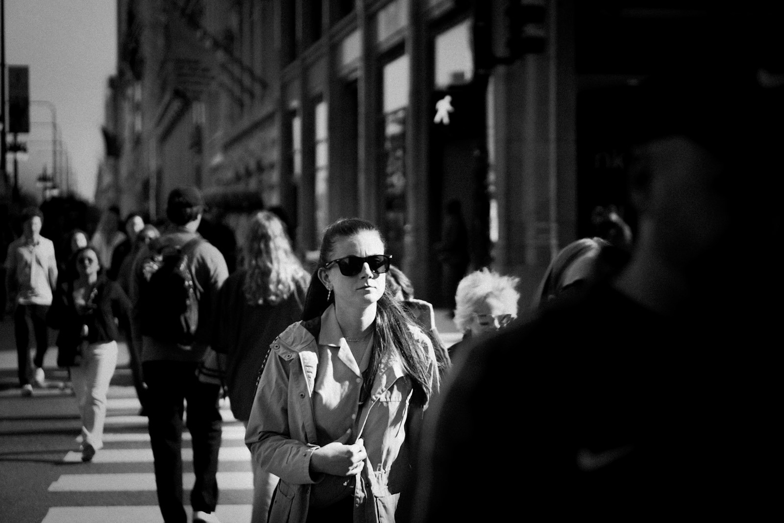 A woman wearing dark Wayfarer-style sunglasses walks across a marked pedestrian crossing in hard sunlight, photographed in black and white, with other pedestrians blurred in front of and behind her.
