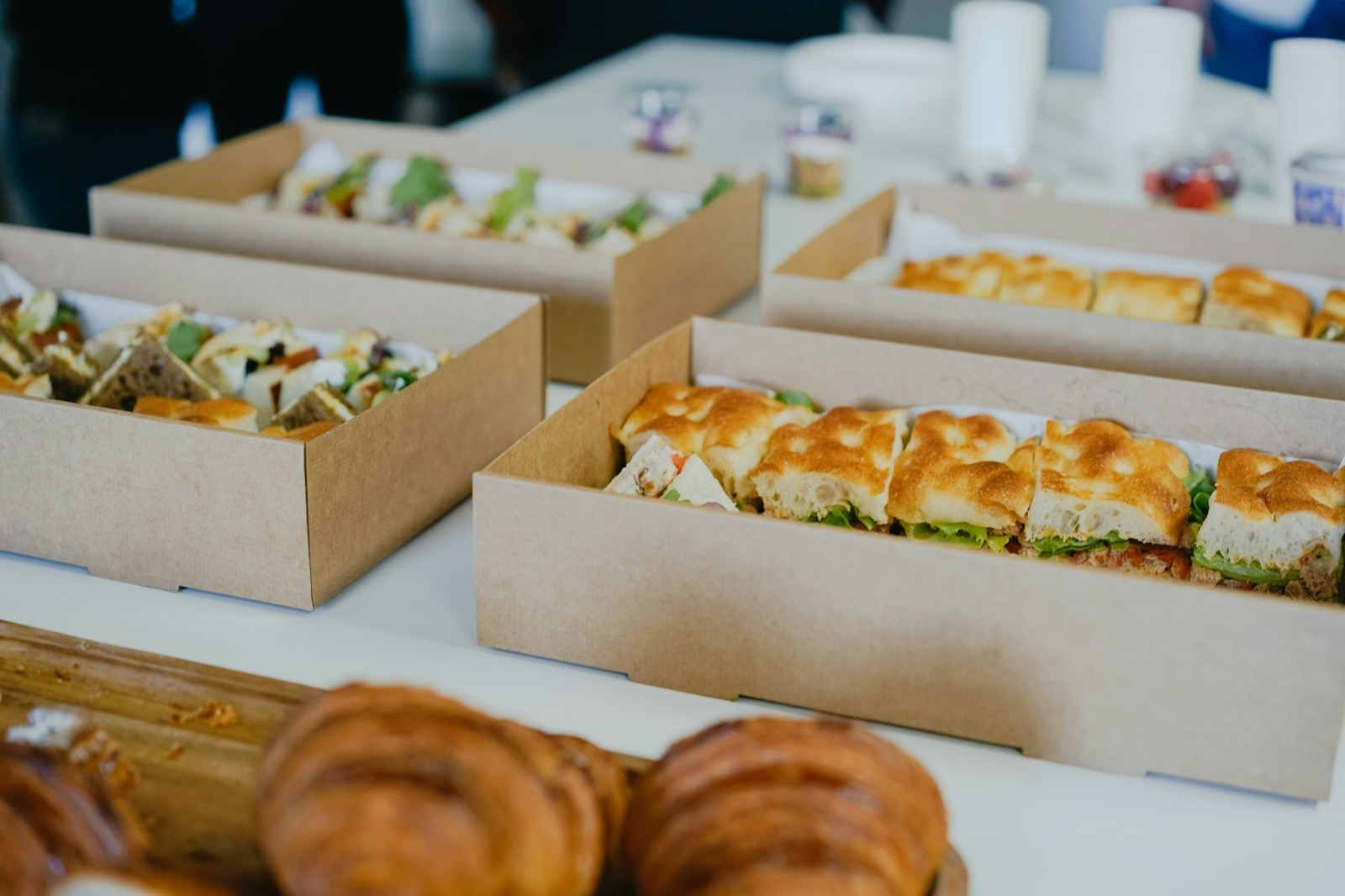 Cardboard catering boxes of focaccia sandwiches and salads on a white table, with croissants in the foreground.