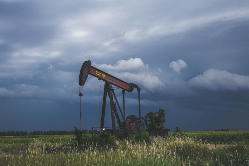 A black and red oil pumpjack stands in a green field under heavy dark storm clouds.