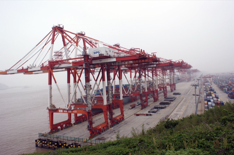 A row of red and white ship-to-shore container cranes along the quay of the Yangshan deep-water port near Shanghai, with stacked containers stretching into the haze on the right.
