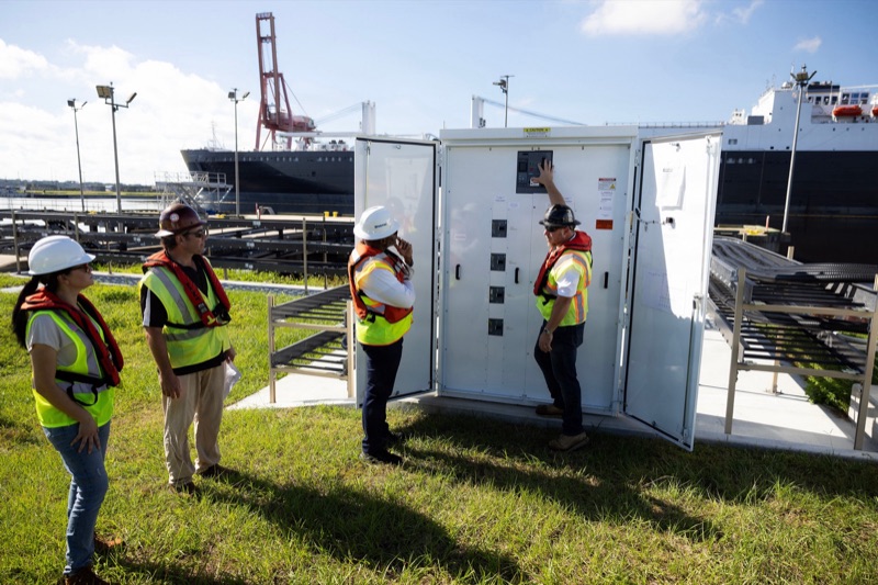 Engineers in hard hats and life vests stand on grass around an open white electrical cabinet on a pier, with a large cargo ship and a red crane behind them.