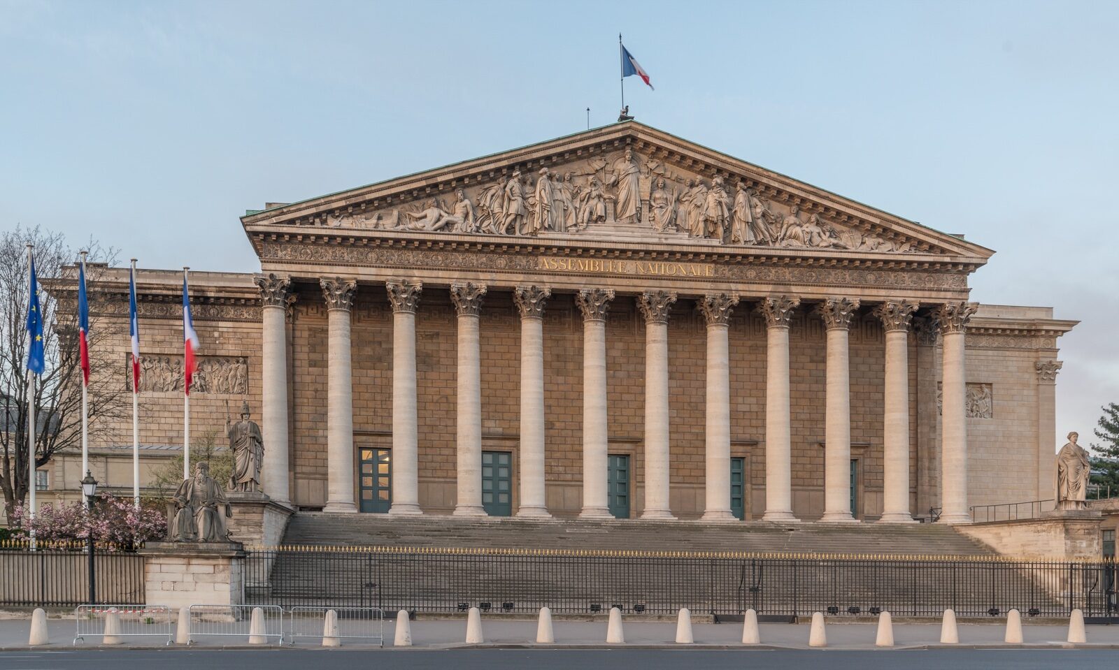 The colonnaded north front of the Palais Bourbon, seat of the French National Assembly, in Paris, with a French flag on the roof and flags along the railings, under a pale sky.