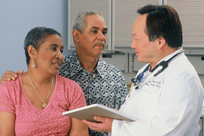 An older woman in a pink top and an older man in a patterned shirt listen as a doctor in a white coat with a stethoscope shows them a clipboard.