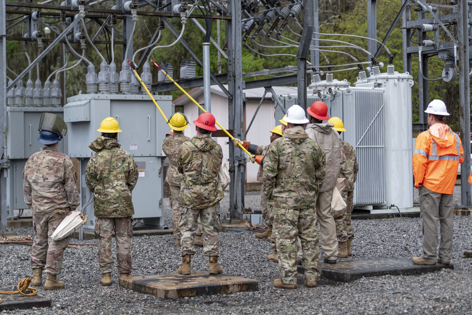 Airmen in hard hats standing in an electrical substation, one reaching up with a long insulated hot stick to operate a switch, with transformers and switchgear behind them.