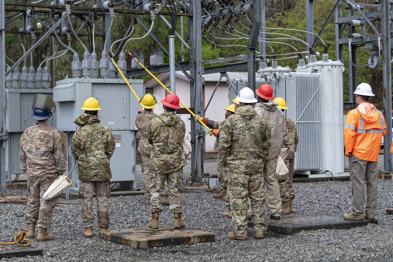 Airmen in hard hats standing in an electrical substation, one reaching up with a long insulated hot stick to operate a switch, with transformers and switchgear behind them.
