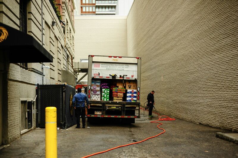 A worker stands at the open back of a delivery truck stacked with boxes of produce in a narrow city alley between brick walls, while another man hoses down the pavement.